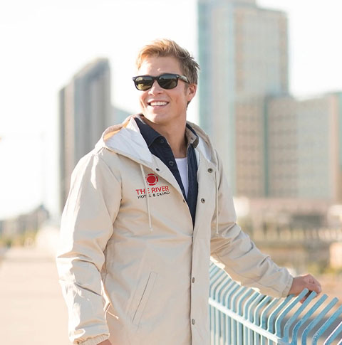 A man in sunglasses smiles outside while wearing a jacket with a custom printed logo on the right chest.
